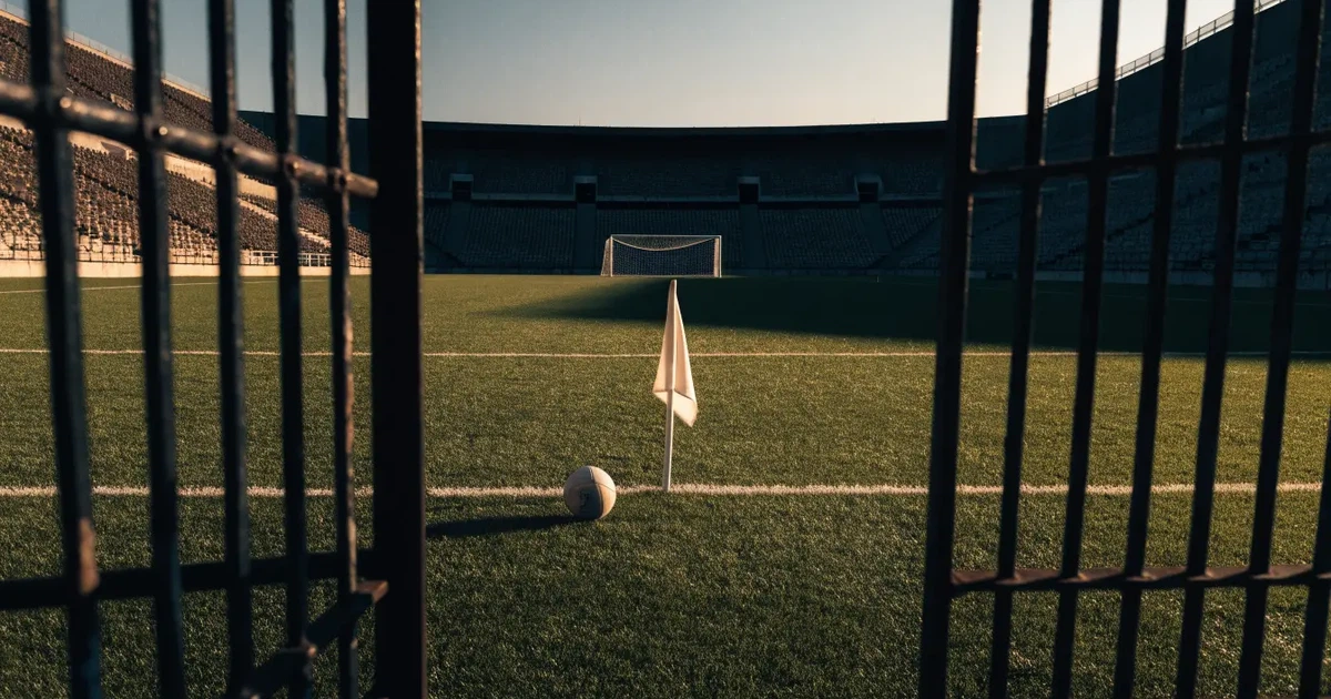 Blick durch ein Stadiontor auf einen grünen Rasen im Abendlicht, ein Ball ruht an der Eckfahne, die Tribünen sind leer.