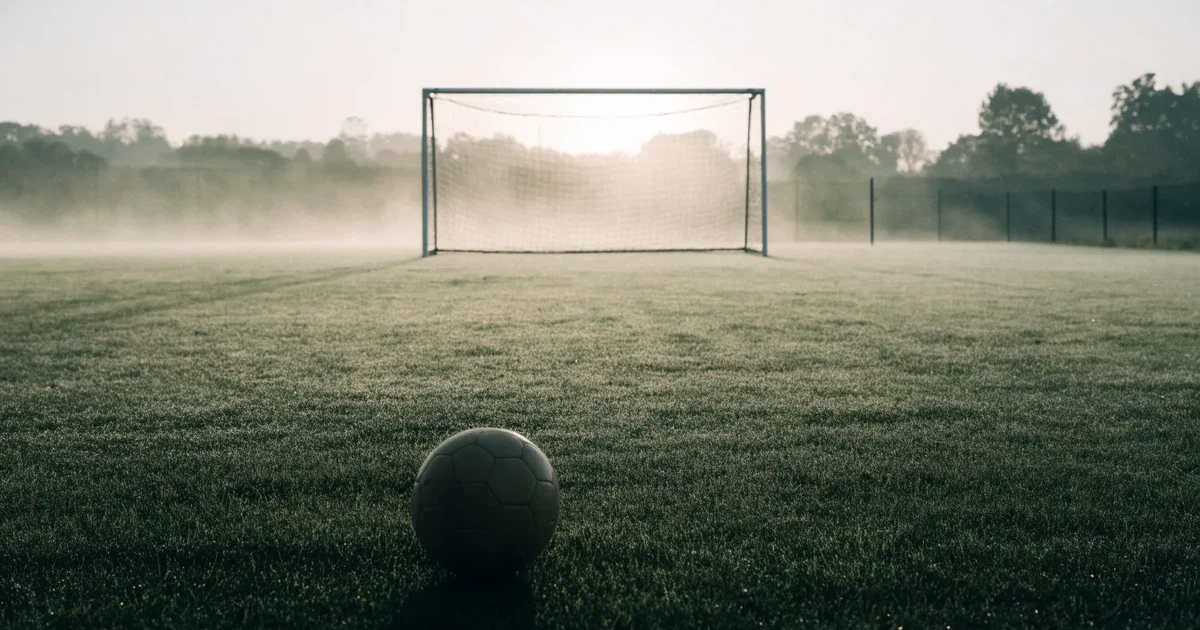Ein Fußballtor mit gespanntem Netz steht auf einem leeren Rasenplatz, davor ein einzelner Ball, Morgendunst liegt über dem Feld.