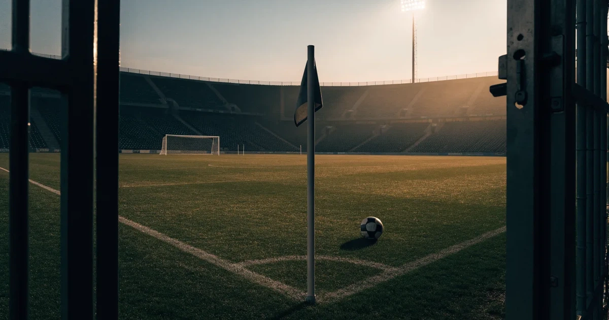 Blick durch ein Stadiontor auf einen grünen Rasen im Abendlicht, ein Ball liegt an der Eckfahne, die Ränge sind leer.