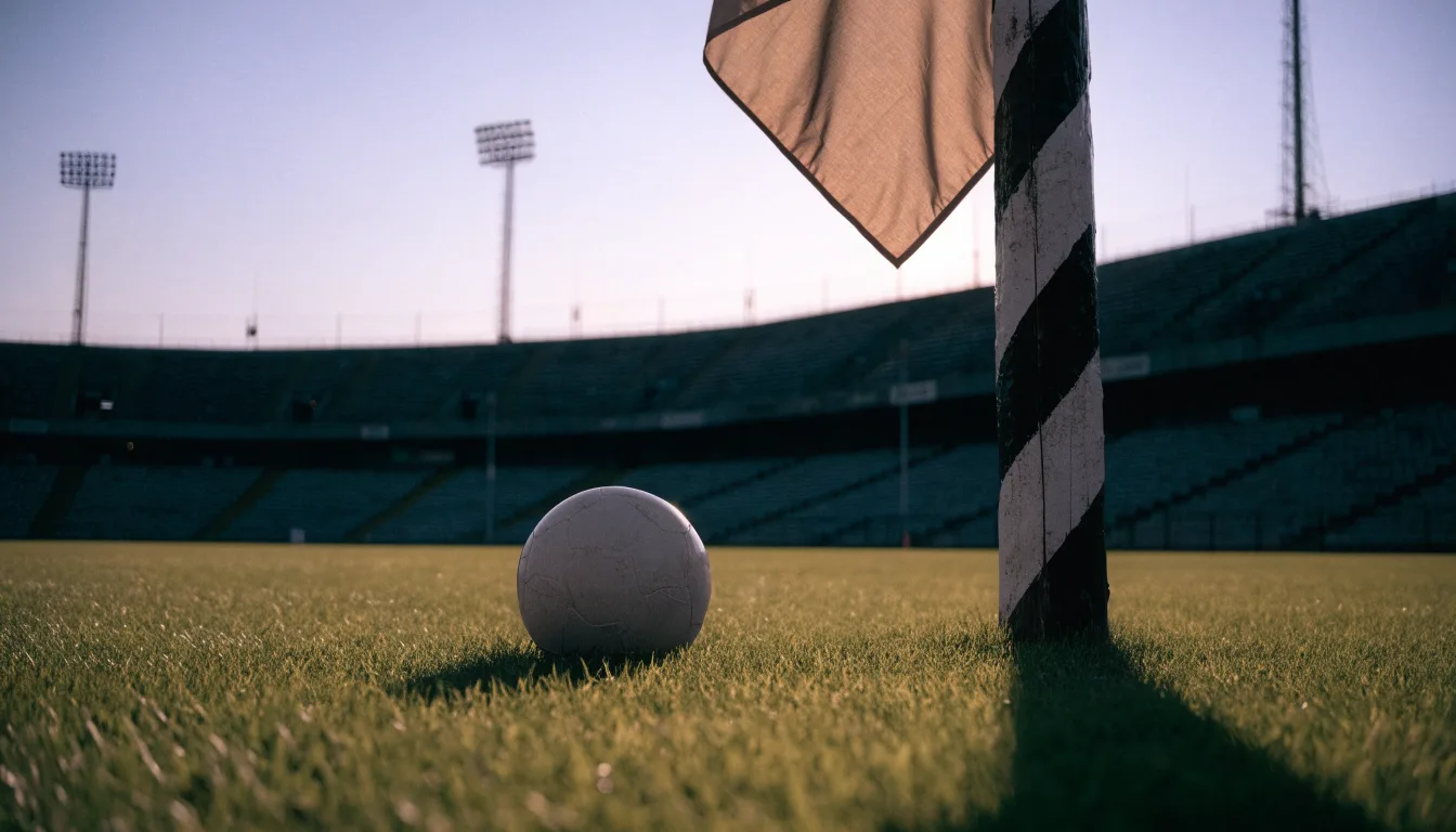 Ein Fußball liegt neben einem Eckfahnen-Pfosten auf einem Rasenplatz, im Hintergrund ein leeres Stadion in der Dämmerung.