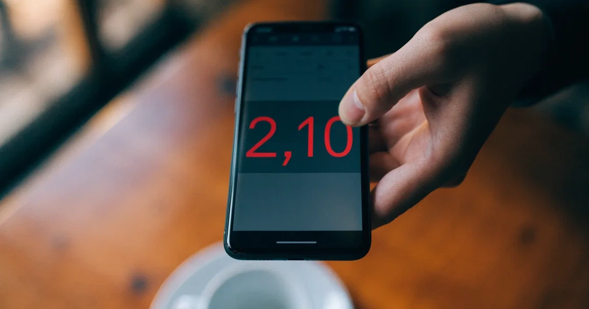 Eine Hand hält ein Smartphone mit dunklem Display, im Hintergrund unscharf ein Café-Tisch mit einer Tasse.
