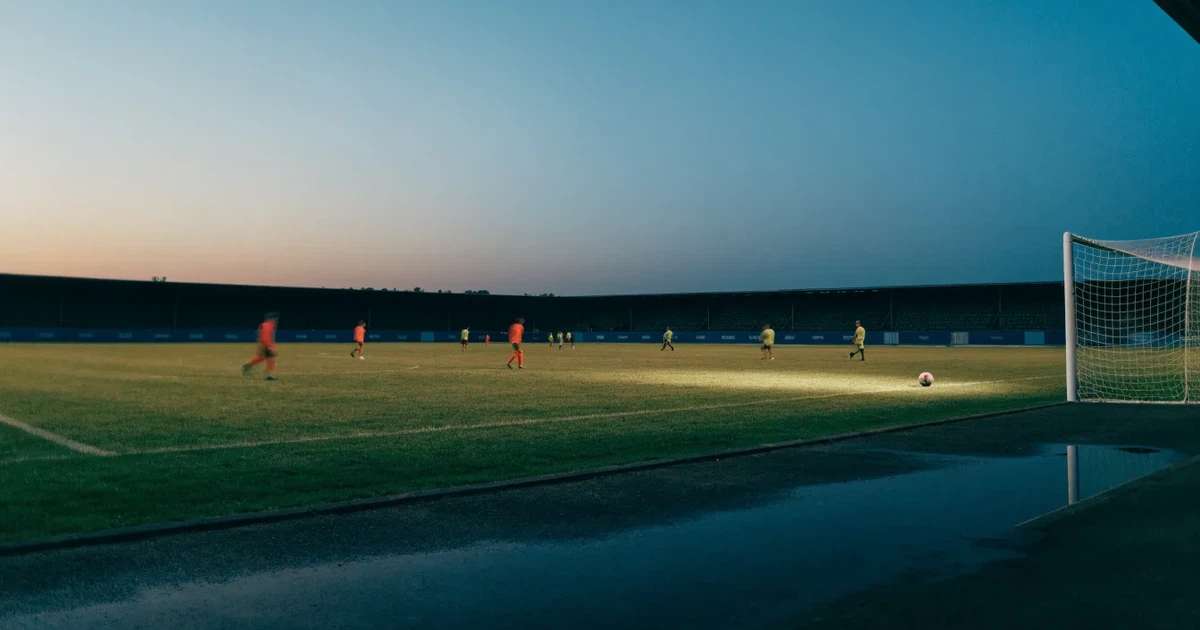 Ein Fußballstadion unter Flutlicht aus großer Entfernung, davor ein dunkler Parkplatz und ein heller Abendhimmel.
