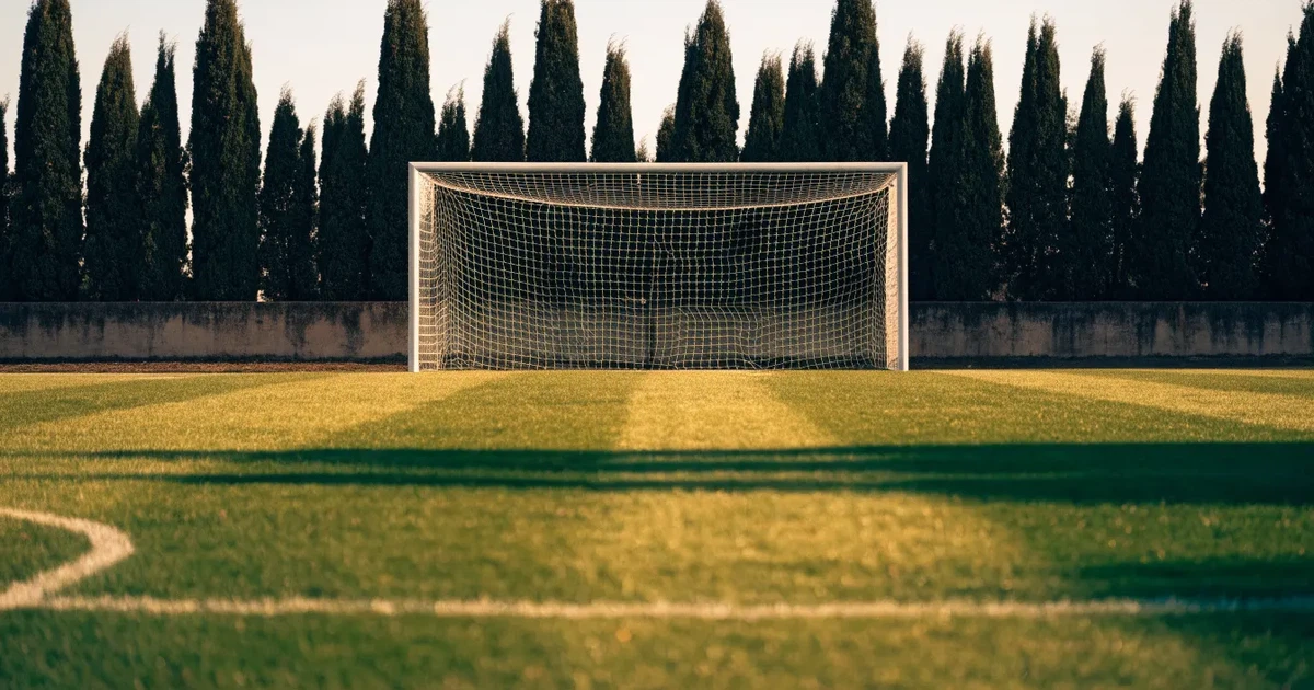 Ein Fußballtor mit Netz auf einem leeren Platz im Abendlicht, der Rasen frisch gemäht, im Hintergrund Zypressen.