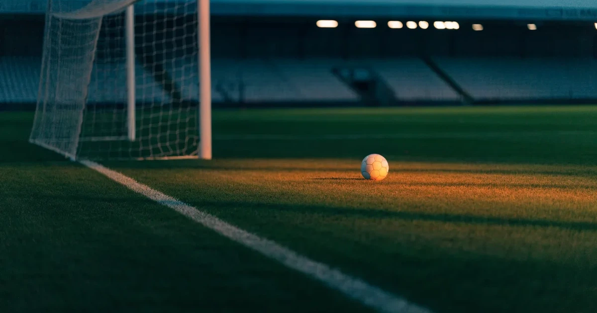 Ein Fußballplatz mit frisch gemähtem Rasen im Abendlicht, ein Ball liegt im Strafraum, im Hintergrund unscharf eine leere Tribüne.