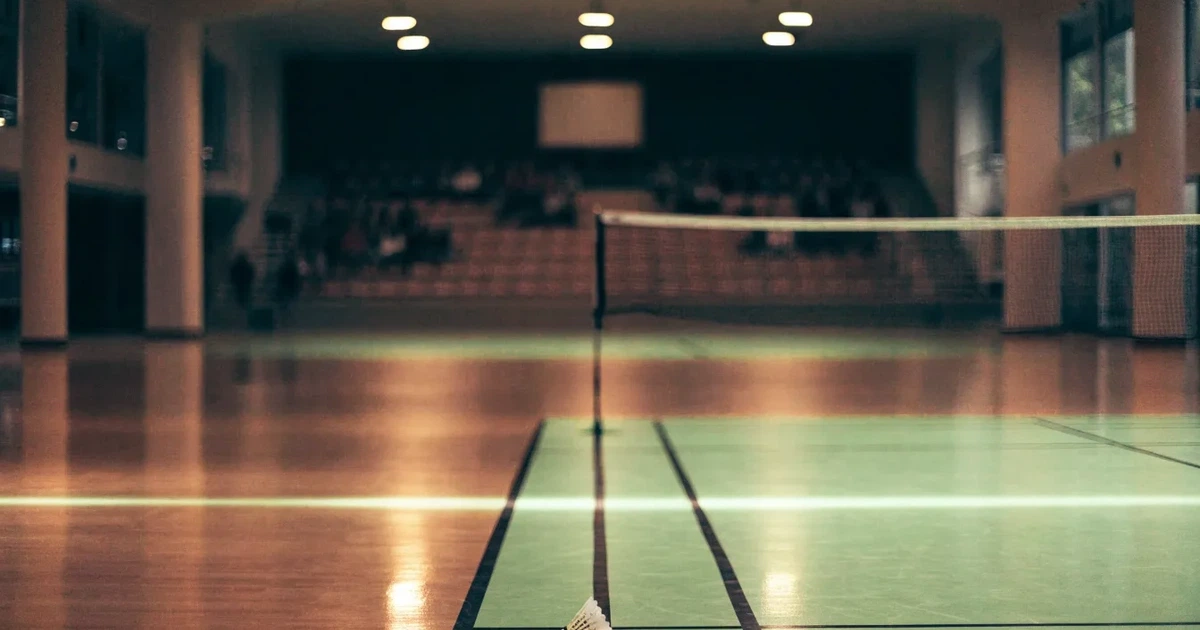 Eine leere Badmintonhalle mit gespanntem Netz und hellen Hallenlichtern, ein Federball liegt auf dem Hallenboden.