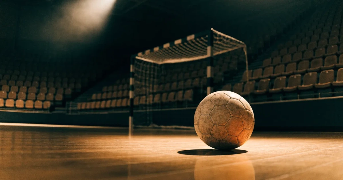 Ein Handball liegt auf dem Hallenboden vor einem Tor mit Netz, die leere Halle wird von Flutlicht beleuchtet.