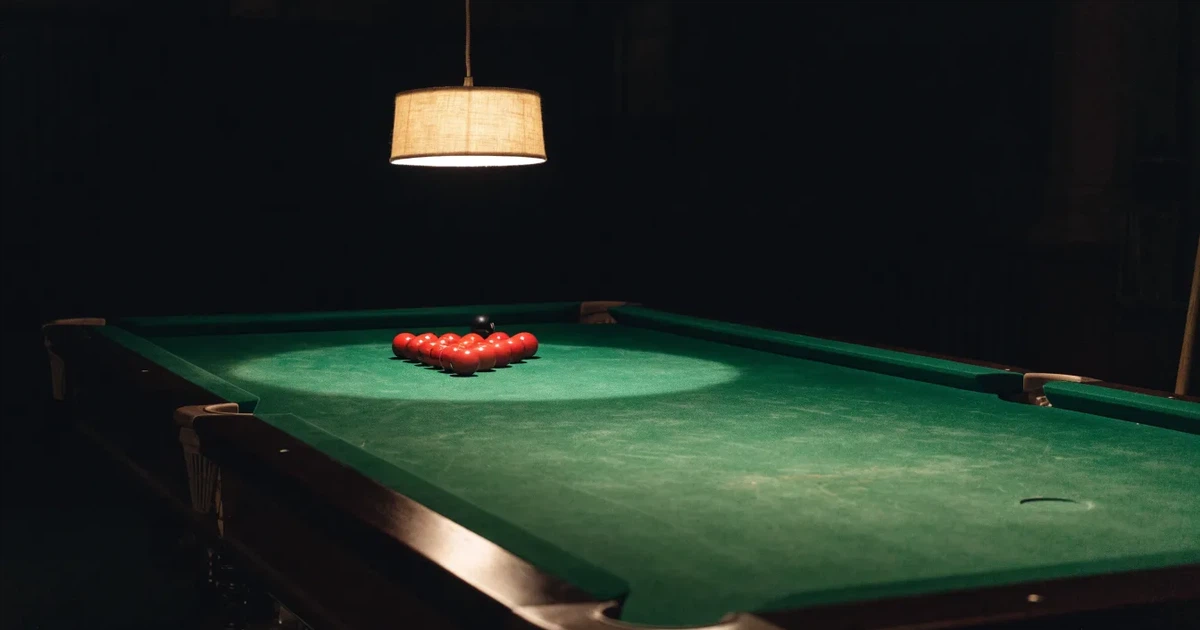 Ein Snookertisch mit grünem Tuch und aufgebauten roten Kugeln in einem abgedunkelten Raum unter einer hängenden Lampe.
