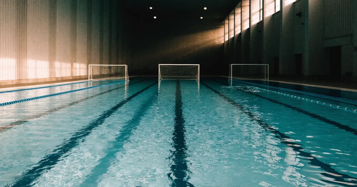 Ein Schwimmbecken mit türkisfarbenem Wasser und zwei schwimmenden Torrahmen am gegenüberliegenden Beckenrand, menschenleer unter Hallenlicht.