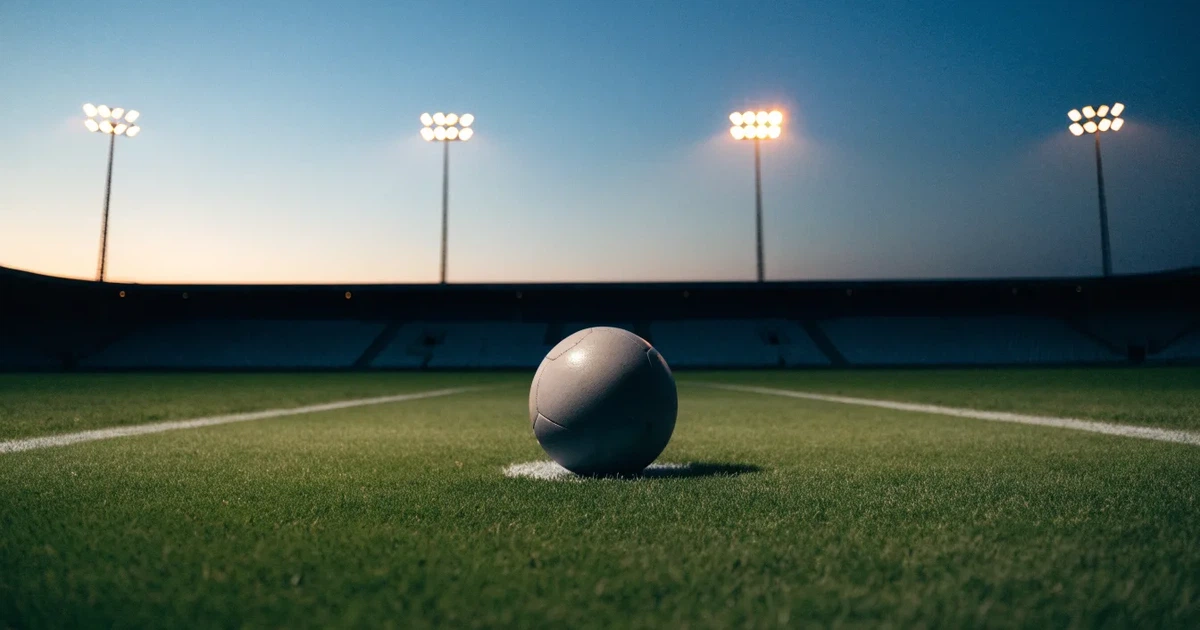 Ein Fußball liegt auf dem Anstoßpunkt eines leeren Rasenplatzes, im Hintergrund eine Tribüne und Flutlichtmasten in der Abenddämmerung.