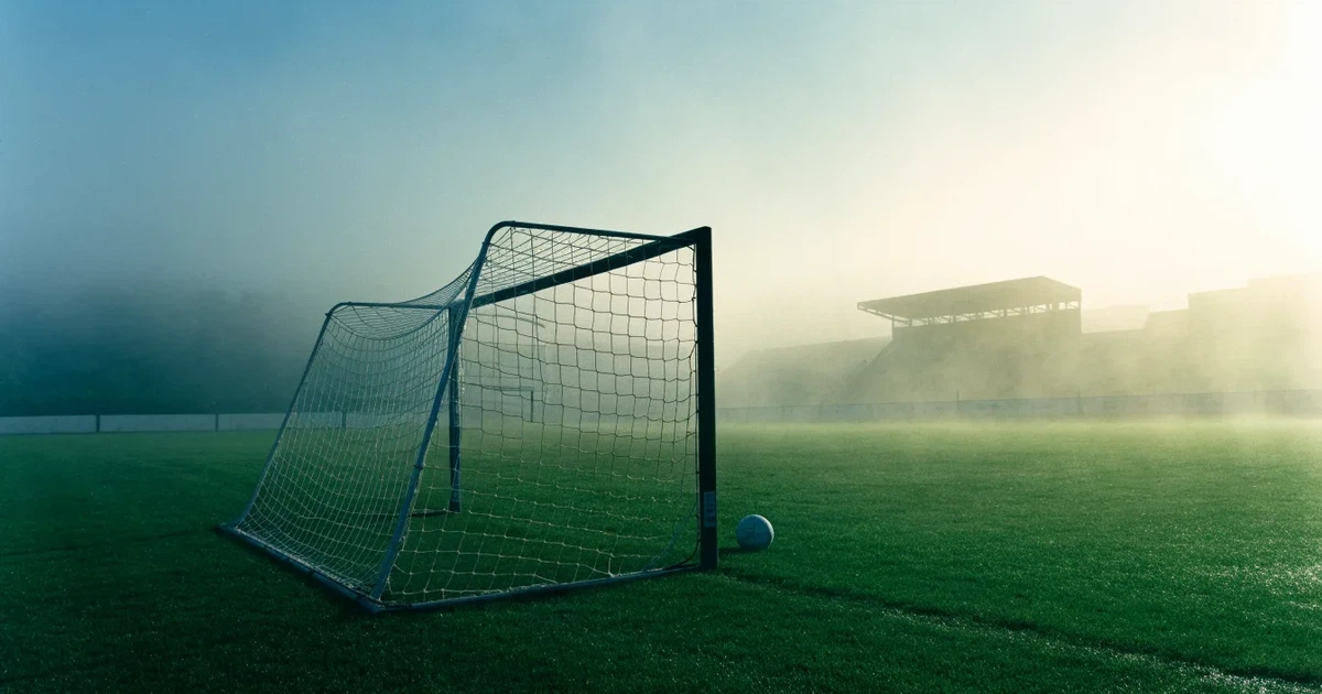 Ein Fußballtor mit Netz steht im Morgennebel auf einem leeren Rasenplatz, im Hintergrund die Silhouette einer kleinen Tribüne.