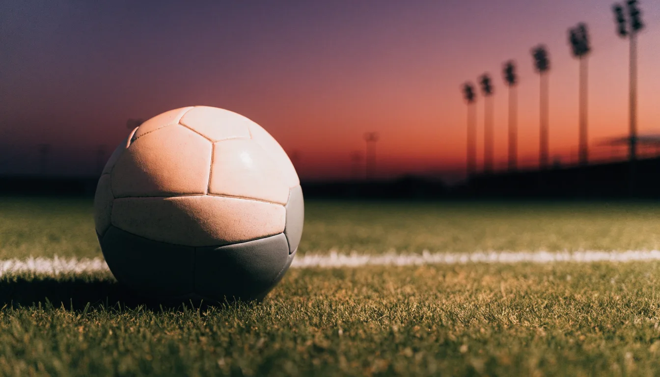 Ein Fußball liegt im Gras eines leeren Platzes, im Hintergrund stehen Flutlichtmasten in der Dämmerung.