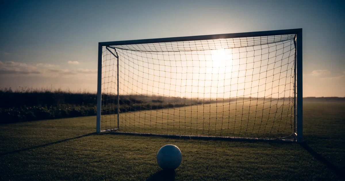 Ein Fußballtor mit Netz im Gegenlicht auf einem leeren Platz am Abend, davor ein Ball auf dem Rasen.