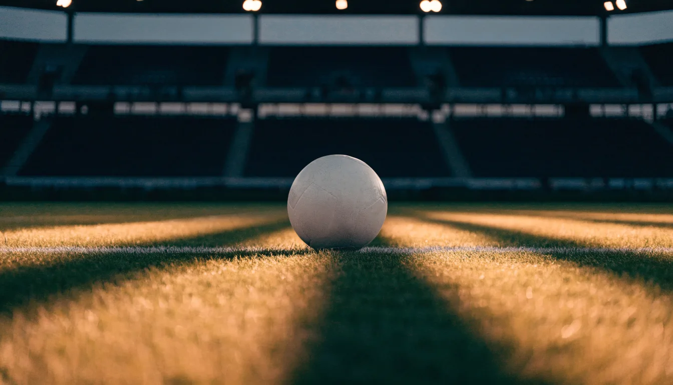 Ein Fußball liegt auf dem Anstoßpunkt eines Rasenplatzes, dahinter eine leere Tribüne im Abendlicht.