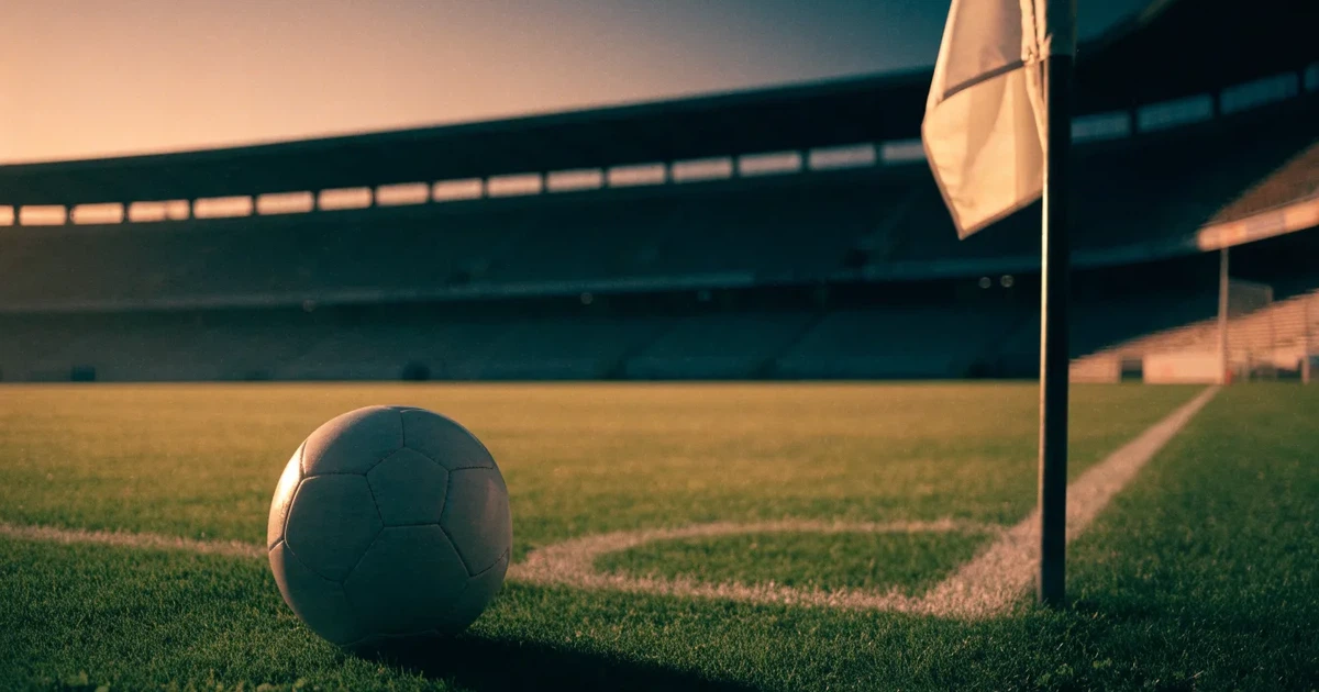 Ein Fußball liegt am Viertelkreis an der Eckfahne eines Rasenplatzes, im Hintergrund leere Tribünen in warmem Abendlicht.