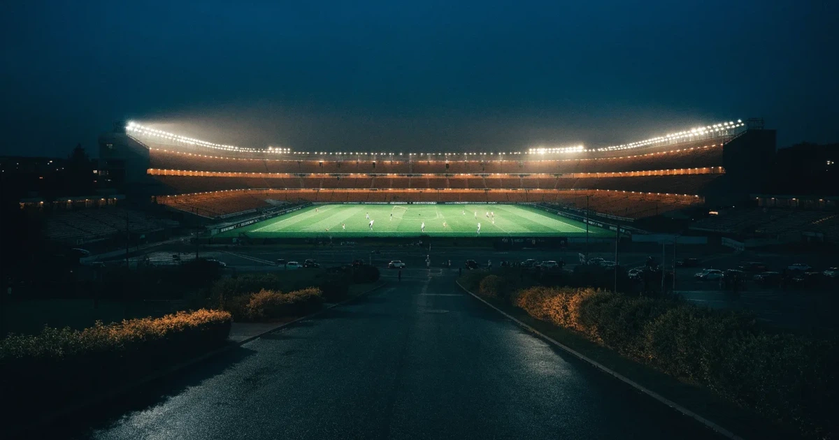 Ein Fußballstadion bei Nacht aus großer Entfernung, hell erleuchtet unter Flutlicht, davor eine dunkle Straße und Bäume.